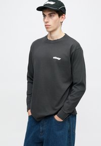 Jovem a usar um boné preto da Obey, uma camisola de mangas longas cinza escuro com o logótipo branco da Obey, e jeans azuis, de pé com as mãos nos bolsos.