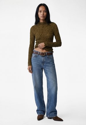 Mujer de pie con una blusa texturizada de manga larga color oliva, vaqueros azules holgados con cinturón marrón y zapatos marrones de punta, sobre fondo blanco.