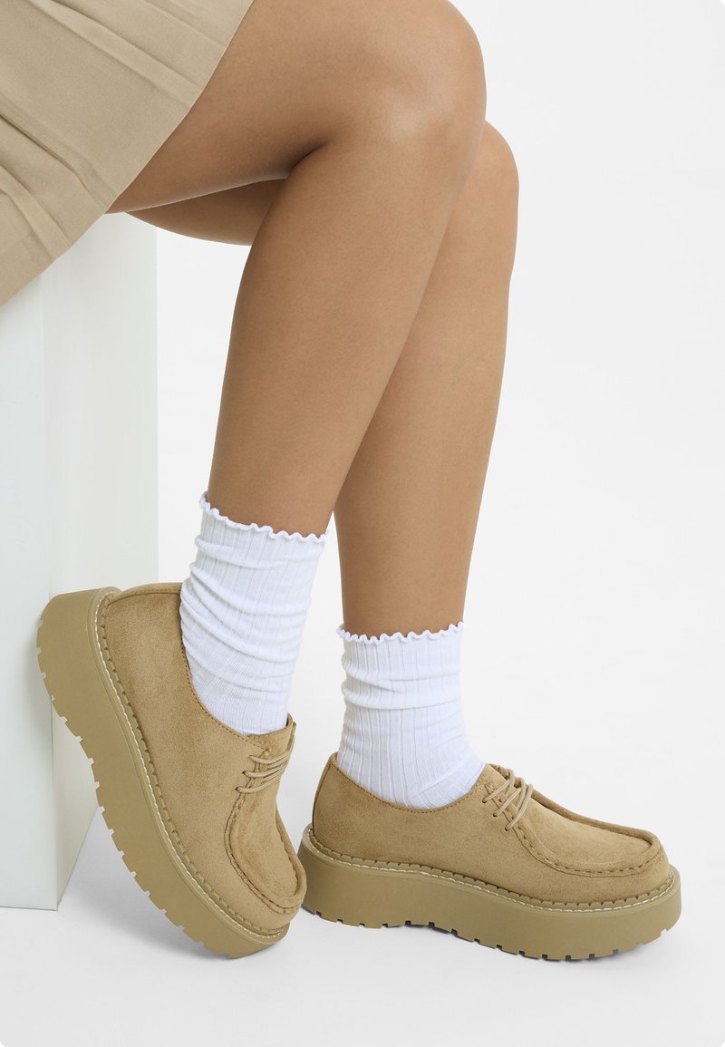 Zapatos de plataforma de ante marrón con puntera redondeada, detalles de cordones y suela de goma gruesa. Se llevan calcetines blancos acanalados con bordes festoneados.
