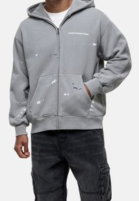 Felpa grigia con zip realizzata in un tessuto morbido, caratterizzata da un logo frontale e dettagli di schizzi di vernice astratti in vari colori, con una vestibilità rilassata e cappuccio.