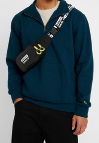 Hombre vestido con sudadera de media cremallera en color azul verdoso y bandolera negra de Adidas con tiradores de cremallera amarillos y una correa con la marca.