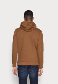 Brown hooded sweatshirt with a soft texture, featuring a kangaroo pocket, ribbed cuffs, and hem. Worn with dark blue jeans.