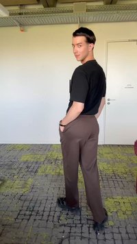 Jeune homme en chemise noire et pantalon marron, les mains dans les poches, regardant par-dessus son épaule dans une pièce avec un tapis à motifs et des murs blancs.