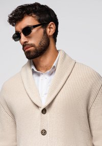Beige gestrickte Strickjacke mit einem Schalkragen, ausgestattet mit drei Knöpfen und einer strukturierten Oberfläche, über einem weißen Hemd mit Kragen getragen. Die Accessoires umfassen schwarze Sonnenbrillen.