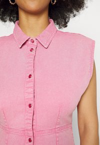 Vaaleanpunainen farkkuasu, jossa on kauluksellinen kaula, lyhyet hihat ja napitus edessä. Huomionarvoista on teksturoitu kangas ja muotoiltu istuvuus.