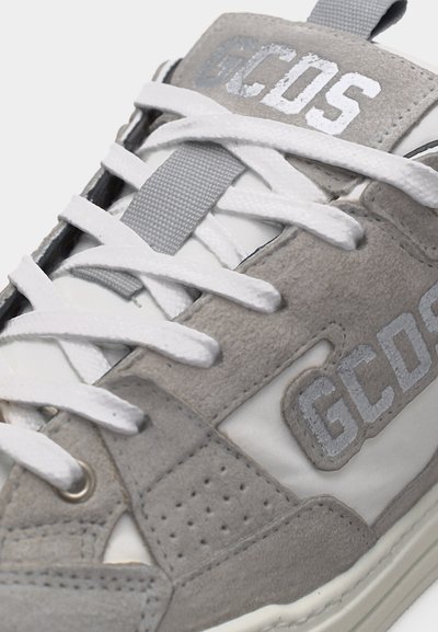 Κοντινή λήψη γκρι και λευκού αθλητικού παπουτσιού με λευκά κορδόνια, σουέντ υφή και το λογότυπο "GCDS" στο πλάι και στην περιοχή της γλώσσας.