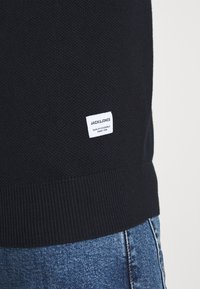 Marinblå stickad tröja med textilerad yta, ribbad nederkant och liten vit etikett med varumärkesnamn och text på undersidan.