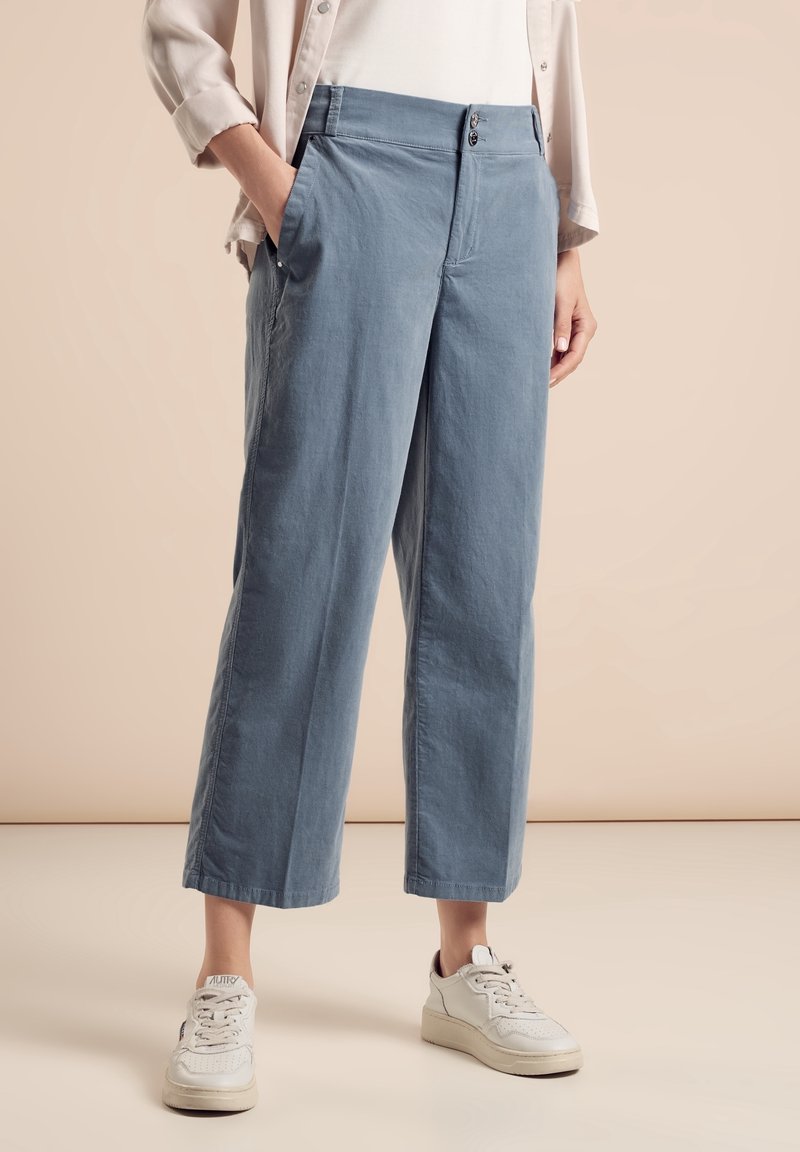 Personne portant un pantalon ample bleu clair court, des baskets blanches et une chemise beige à manches longues, les mains dans les poches.