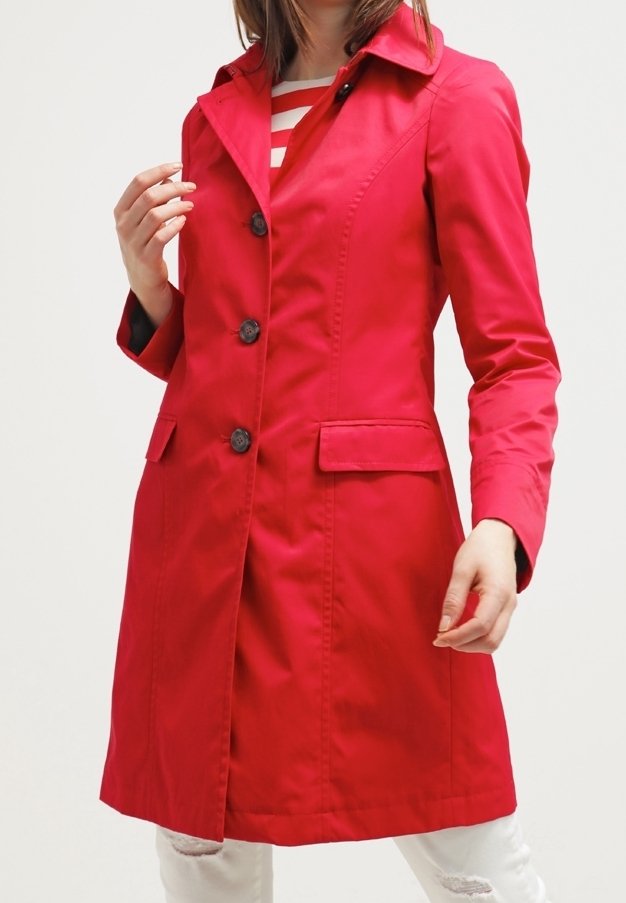Femme portant un manteau rouge mi-long avec des boutons noirs sur une chemise rayée rouge et blanche et un jean blanc déchiré, sur un fond uni.