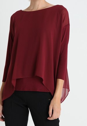 Vrouw draagt een losse, gelaagde kastanjebruine top met lange mouwen en een asymmetrische zoom, gecombineerd met een zwarte broek.