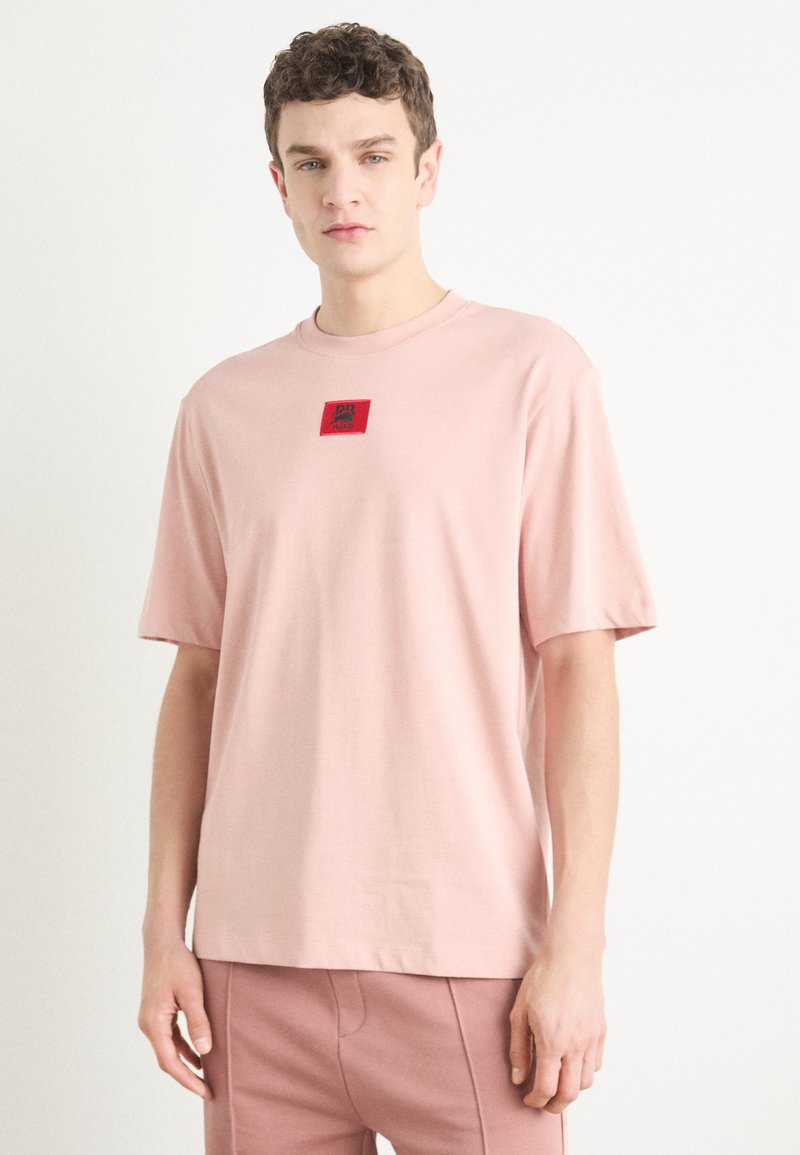 T-shirt en coton rose clair avec un col rond, des manches courtes et un patch logo rouge carré sur la poitrine. Texture lisse et coupe décontractée.