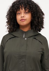 Olivgrön huva-jacka i lättviktsmaterial med dragkedja fram, tryckknappsstängning och minimalistisk design med rundade axelsömmar.