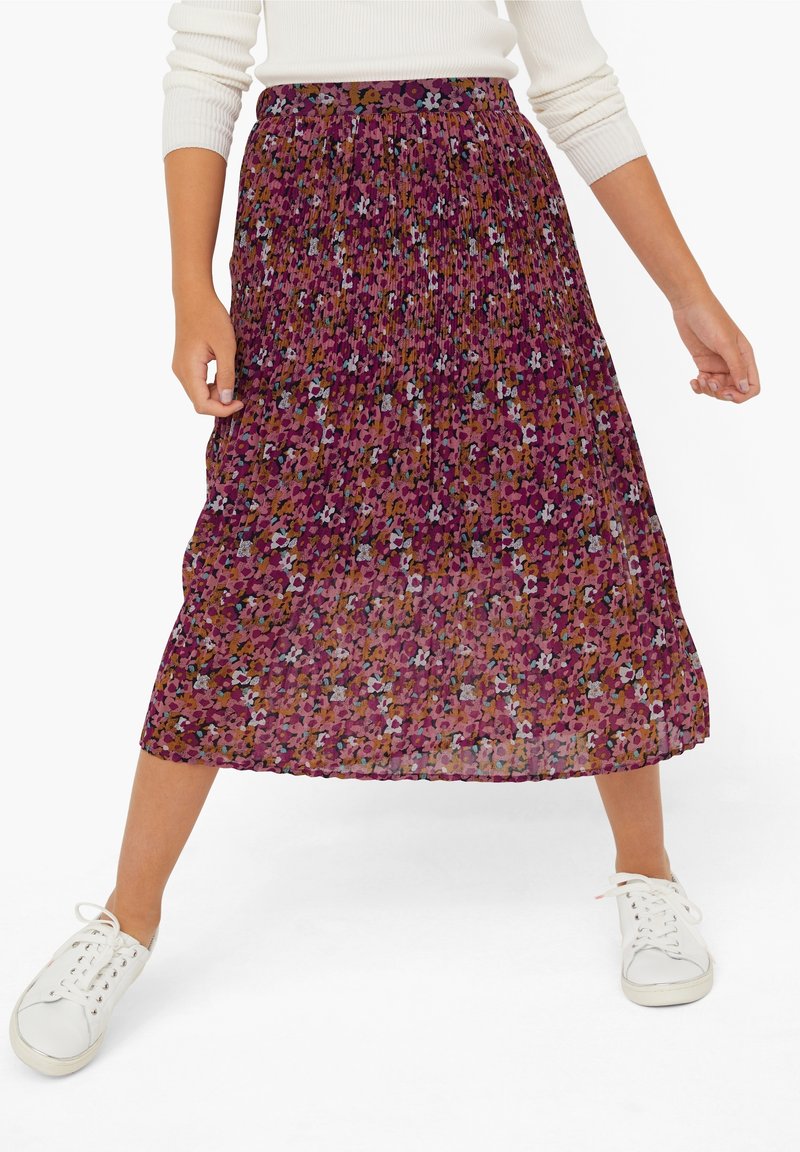 Femme portant une jupe midi plissée à motif floral coloré, associée à un pull blanc et des baskets blanches.