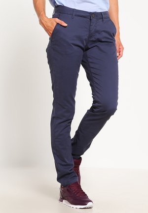 Osoba ubrana w granatowe, dopasowane spodnie i bordowe trampki, stojąca z jedną ręką w kieszeni na jednolitym tle.