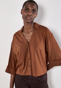 Brun kortærmet skjorte med knapper og krave, der har en afslappet pasform og glat tekstur. Subtil glans og detalje med frontlomme.
