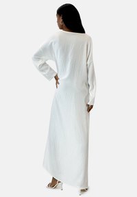 Robe blanche longue et ample en tissu texturé, avec des manches larges et un décolleté rond. L'ourlet est légèrement plus long à l'arrière.