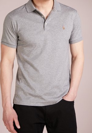 Mężczyzna w szarej koszulce polo z krótkim rękawem z małym haftowanym logo i czarnych spodniach, z ręką w kieszeni, na neutralnym tle.