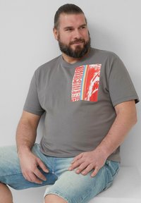 Graues Kurzarm-T-Shirt mit einem rot-weißen Grafikdesign auf der linken Brust. Glatte Stoffqualität, lässiger Schnitt, kombiniert mit hellen Denim-Shorts.