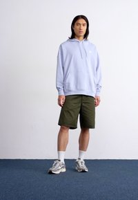 A young person stands in a light blue hoodie and green shorts on a blue carpet, with a plain white background.