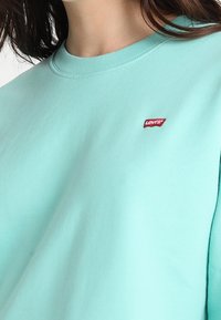 Donna che indossa un maglione leggero color turchese con un piccolo logo rosso di Levi's cucito sulla parte superiore sinistra del petto.