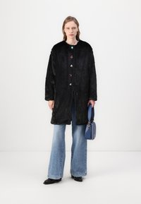 Črn plašč iz umetnega krzna z dolgimi rokavi in pisanimi cvetličnimi gumbi, okroglim ovratnikom in stranskimi žepi. Obučen preko razširjenih kavbojk in v kombinaciji z modro torbico.