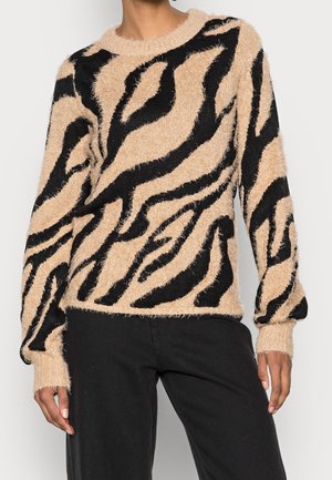 Person iført beige og sort fluffy sweater med abstrakt zebra-lignende mønster og sorte bukser, stående mod en ensfarvet baggrund.