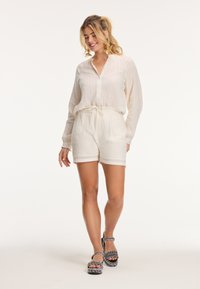 Blusa ligera blanca de manga larga con botones en la parte delantera, combinada con pantalones cortos crema que tienen una cinturilla elástica y una textura sutil. Sandalias de patrón en blanco y negro.