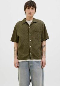 Korte mouwen, olijfgroene shirt met een gestructureerde stof, voorzien van een knoopsluiting aan de voorkant en een borstzak, gecombineerd met lichtblauwe jeans.