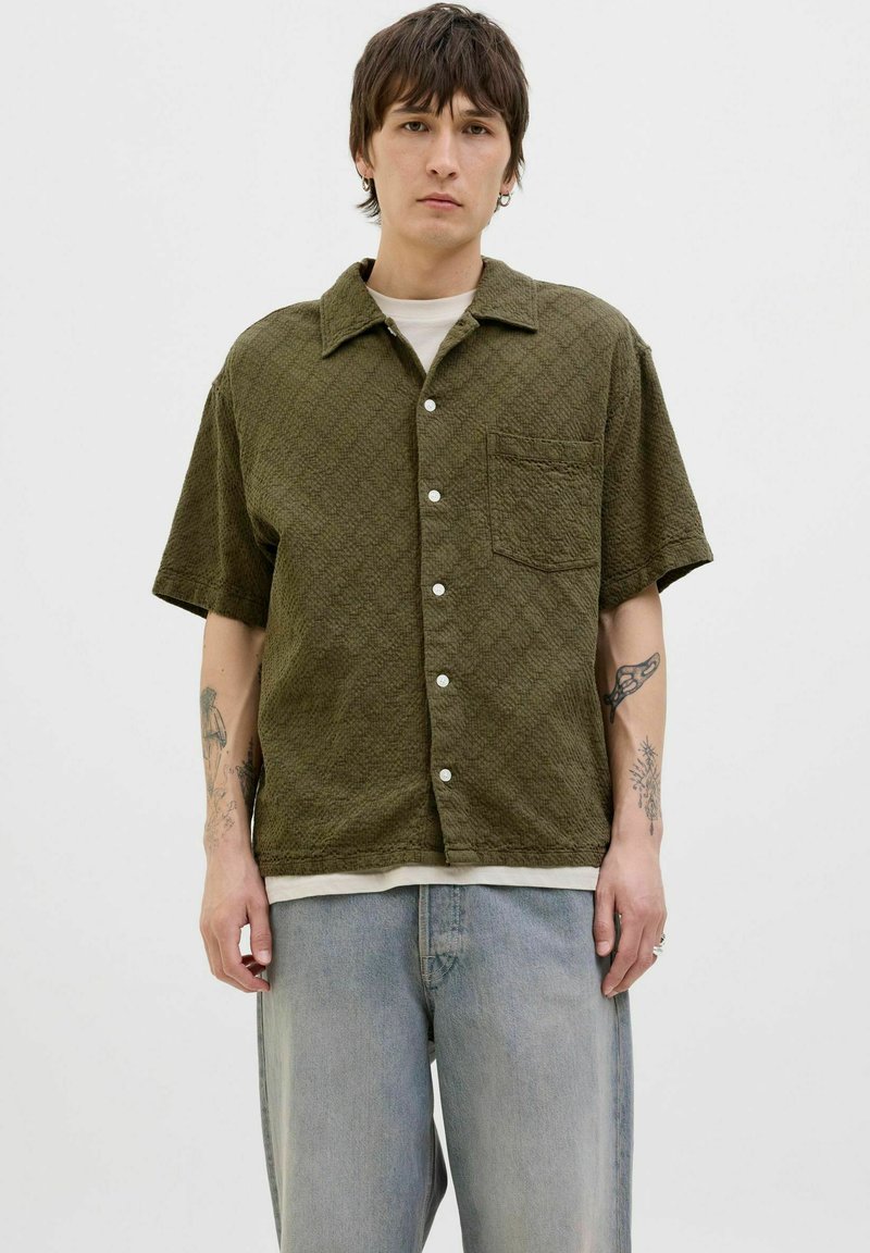 Korte mouwen, olijfgroene shirt met een gestructureerde stof, voorzien van een knoopsluiting aan de voorkant en een borstzak, gecombineerd met lichtblauwe jeans.
