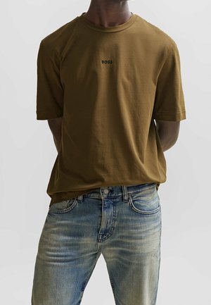 Homme portant un t-shirt marron BOSS à manches courtes et un jean bleu délavé, mains placées derrière le dos, fond neutre.