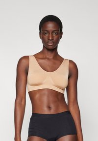 Nude pull-on bralette dengan tali lebar dan tekstur halus, menampilkan desain tanpa jahitan dan tanpa perangkat keras yang terlihat. Dipasangkan dengan celana dalam hitam dengan pinggang tinggi.