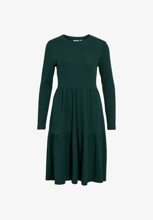 Robe à manches longues, de couleur vert foncé, avec un corsage ajusté et une jupe évasée à volants. Fabriquée en tissu côtelé. Encolure ronde et sans embellissements supplémentaires.