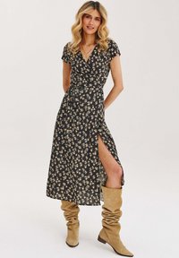 Čierna kvetinová zavinovacia šaty s krátkymi rukávmi, bočným rozparkom a viazaním v páse. Skombinované s béžovými vysokými čižmami. Hladká textúra látky.