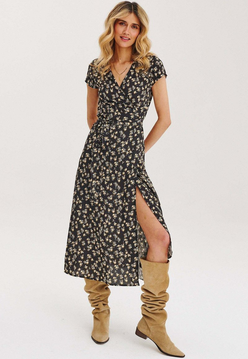 Čierna kvetinová zavinovacia šaty s krátkymi rukávmi, bočným rozparkom a viazaním v páse. Skombinované s béžovými vysokými čižmami. Hladká textúra látky.