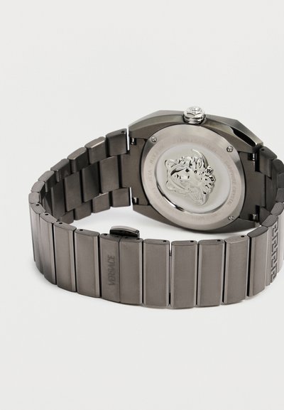 Montre-bracelet en métal noir avec un bracelet à maillons, emblème de Méduse gravé au dos, et le nom de marque "Versace" sur la boucle.