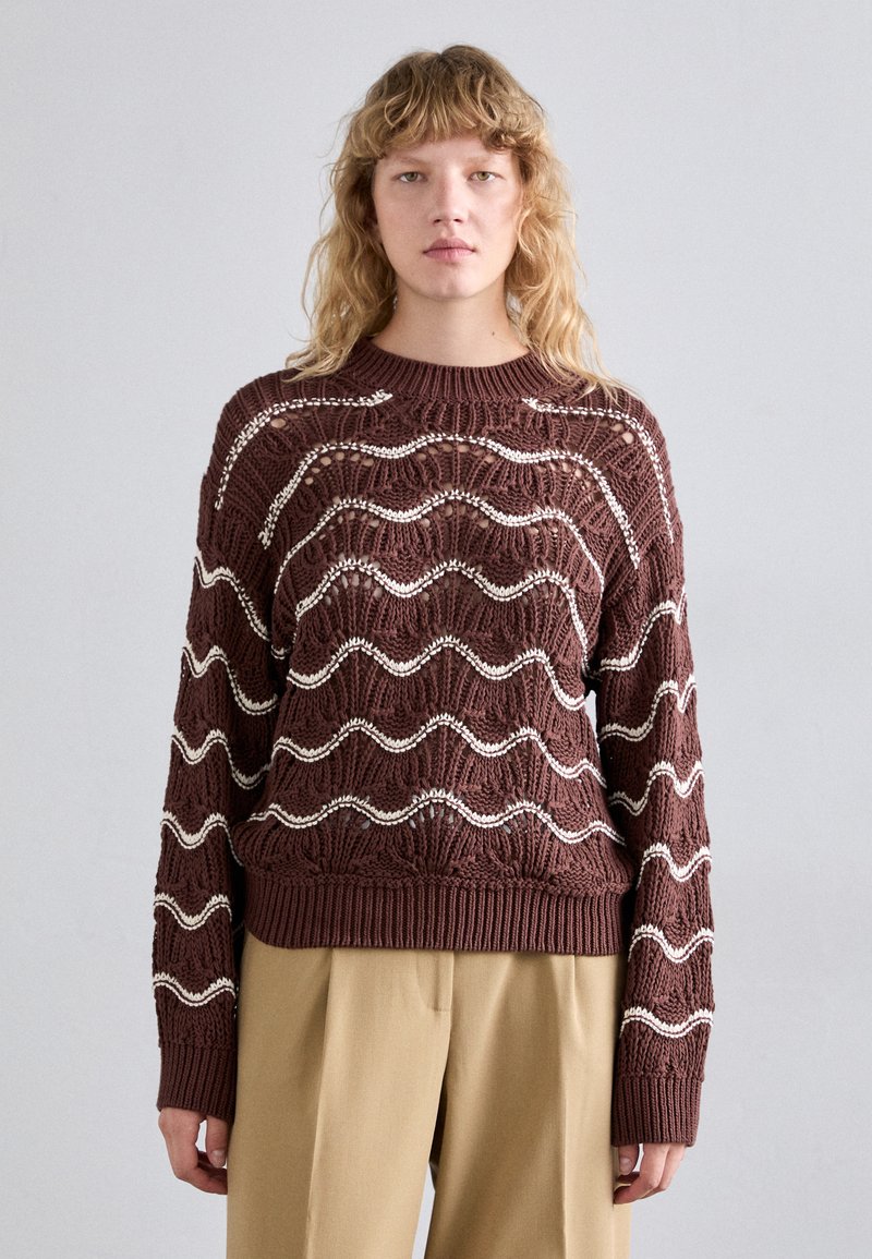 Brun strikket sweater med et bølge mønster i hvid, ribbede manchetter og kant, rund hals og en løs pasform. Sammen med beige bukser.