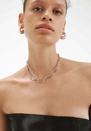 Collar de cadena de plata con eslabones entrelazados de diferentes formas, llevado en un cuello desnudo. Pendientes simples complementan el collar.