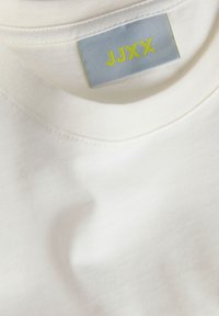 Κοντινή λήψη ενός λευκού T-shirt με στρογγυλή λαιμόκοψη που δείχνει μια γκρι ετικέτα με κίτρινο κείμενο "JJXX" ραμμένη μέσα στο γιακά.