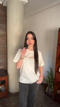 Νεαρή γυναίκα με μακριά σκούρα μαλλιά, φοράει άσπρο μπλουζάκι και μαύρο παντελόνι, κρατάει μικρό μικρόφωνο σε φωτεινό δωμάτιο με ξύλινα έπιπλα και φυτό.