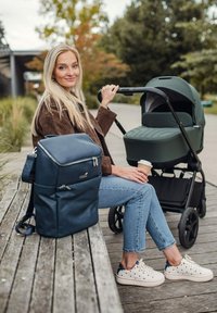 Tmavě modrý batoh s texturovaným materiálem, hnědá bavlněná bunda, světle modré džíny, bílé tenisky s tmavě modrými puntíky a zelený kočárek.