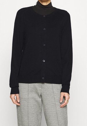 Cárdigan negro con mangas largas y un cuello redondo, que cuenta con seis botones en el frente. Llevado sobre un cuello alto oscuro y pantalones con estampado.
