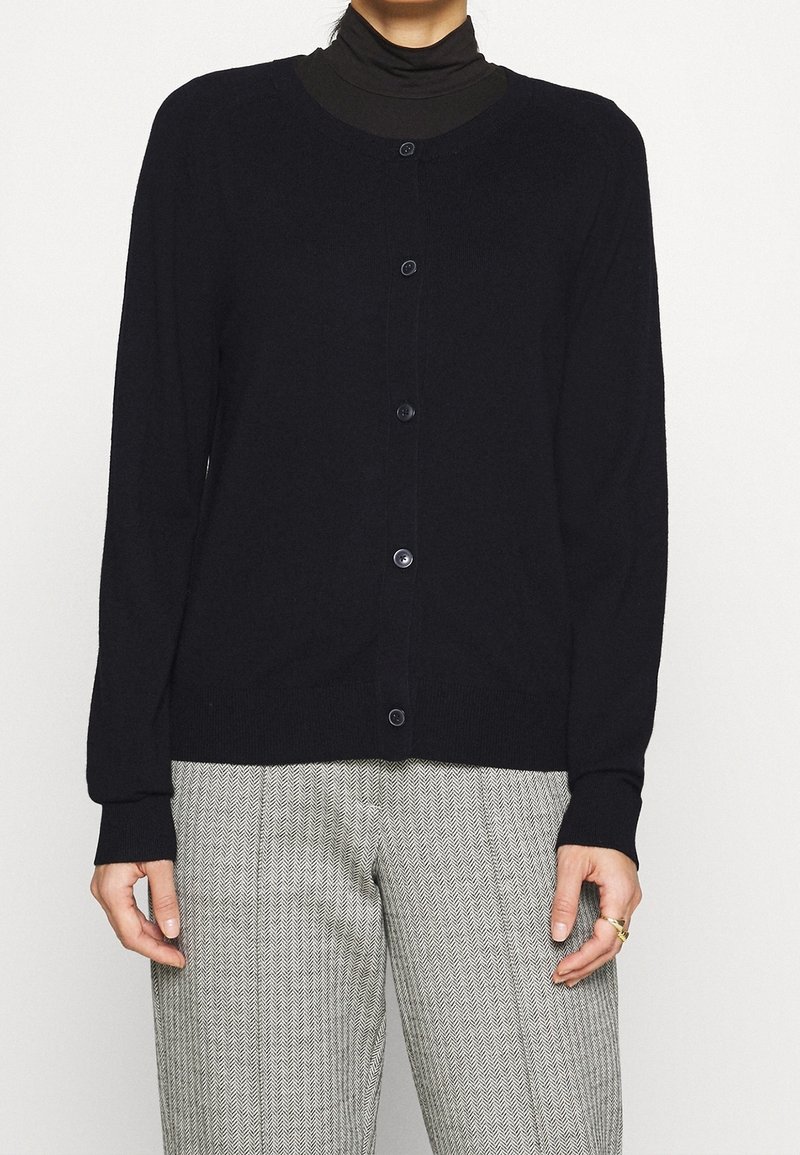 Cárdigan negro con mangas largas y un cuello redondo, que cuenta con seis botones en el frente. Llevado sobre un cuello alto oscuro y pantalones con estampado.