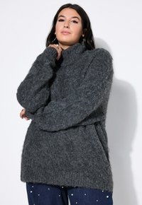 Grau oversized Strickpullover mit hohem Kragen, aus strukturiertem, fluffigem Material und mit überschnittenen Schultern, kombiniert mit dunkelblauen Jeans, die mit Perlen verziert sind.