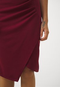 Robe bordeaux longueur genou avec un design ajusté, des fronces sur les côtés et un ourlet asymétrique. Fabriquée à partir d'un tissu lisse et extensible.