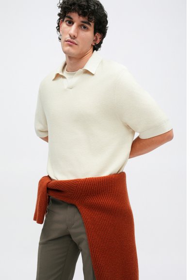 Pleteni polo majica v kremni barvi z teksturiranim vzorcem; olivno zeleni hlače; oversize rjava jopica zavezana okoli pasu.