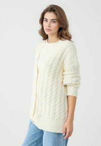 Cardigan a maglia a trecce crema con vestibilità oversize, dettagli in perle e polsini a coste. Indossato sopra una blusa leggera e abbinato a jeans blu.