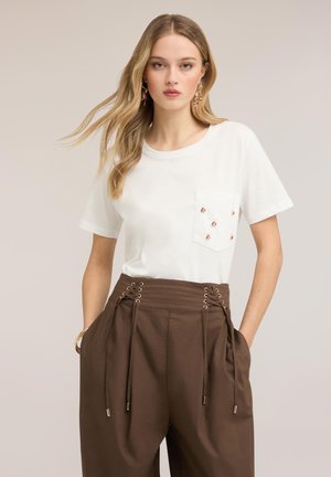 Jonge vrouw met lang blond haar die een wit T-shirt draagt met kleine versieringen op de zak en een hoog getailleerde bruine broek met veterdetail.