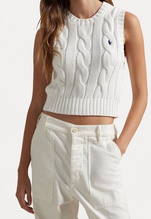 Femme porte un pull court blanc sans manches en maille torsadée et un pantalon blanc taille haute avec la main dans la poche sur fond uni.