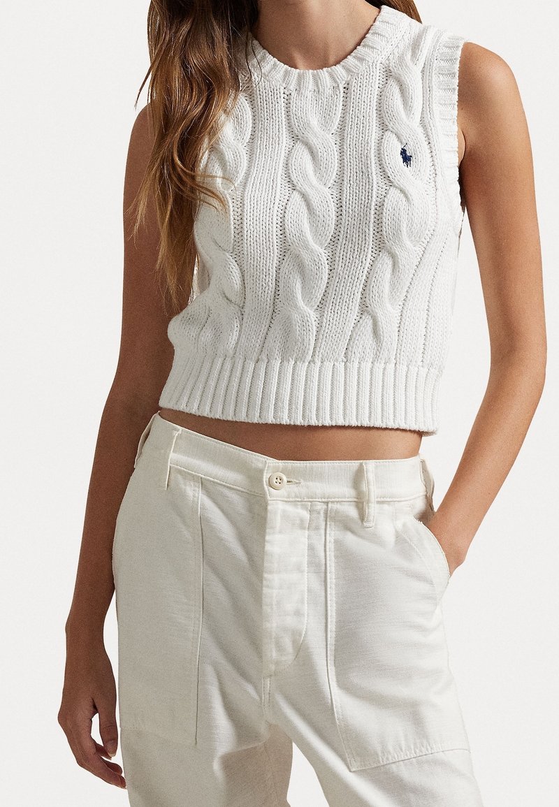 Femme porte un pull court blanc sans manches en maille torsadée et un pantalon blanc taille haute avec la main dans la poche sur fond uni.