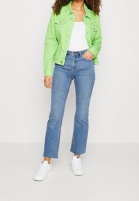 Chaqueta de denim verde brillante con cierres de botones, llevada sobre una camiseta blanca. Combinada con jeans acampanados de lavado claro y zapatillas blancas.